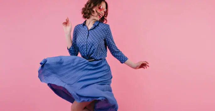 a woman in a blue skirt and polka dot shirt is dancing on a pink background .