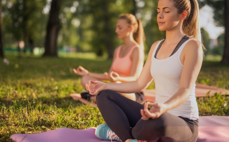 two women sit on yoga mats in a park meditating