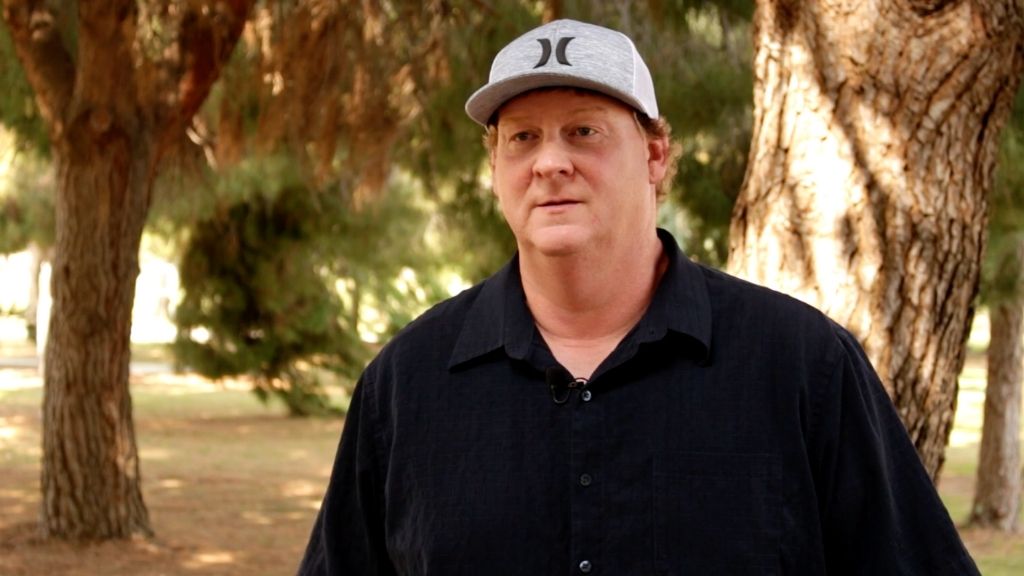 a man wearing a hurley hat and a black polo stands in front of trees