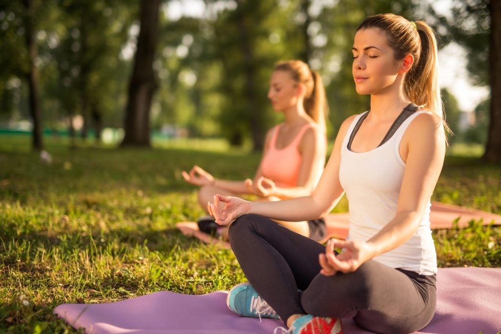 two women sit on yoga mats in a park with their eyes closed meditating